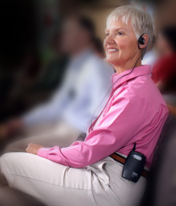 Una mujer, vestida con una camisa rosa y pantalones caquis, sonríe mientras usa un dispositivo de audición inalámbrico llamado "sistema FM".