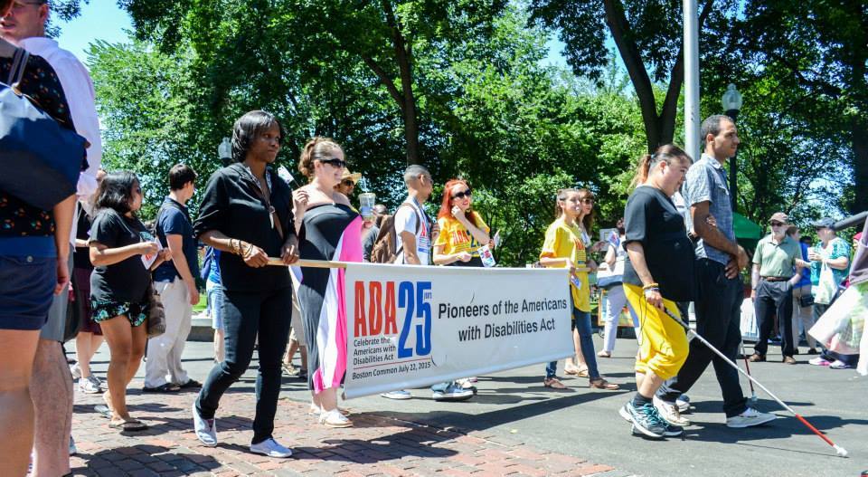 People march in the ADA Anniversary parade holding a banner reading ADA 25 Pioneers of the Americans with Disabilities Act