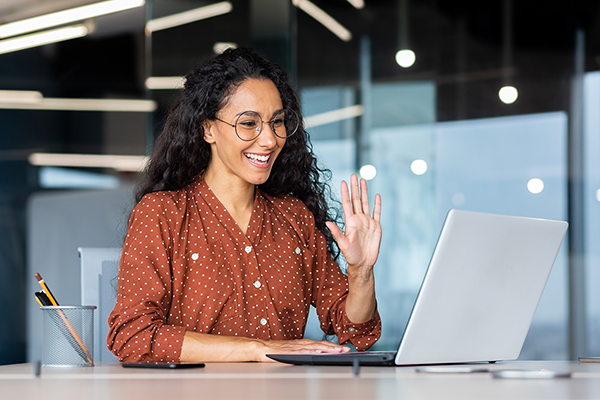 A medium-skinned brunette woman, waving at a silver laptop in a modern office. She is wearing round black, circular glasses, and an orange and white spotted shirt.