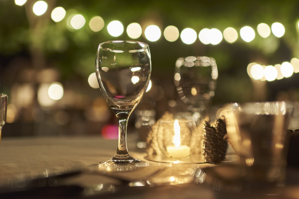 A nighttime outdoor celebration at a restaurant. There are string lights in the background, a table with glasses and a candle.