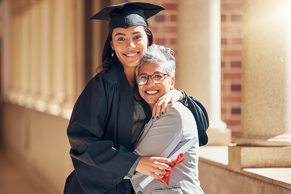 A medium-skinned younger woman and medium-skinned older woman standing and smiling together. The younger woman is wearing black graduation robes.