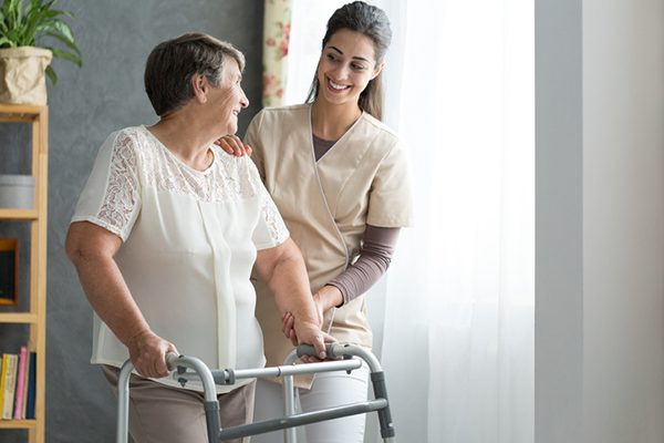 A light-skinned woman with short grey-brown hair. She is using a walker. A smiling medical professional in tan scrubs assists her by placing a steadying hand on her shoulder and forearm.