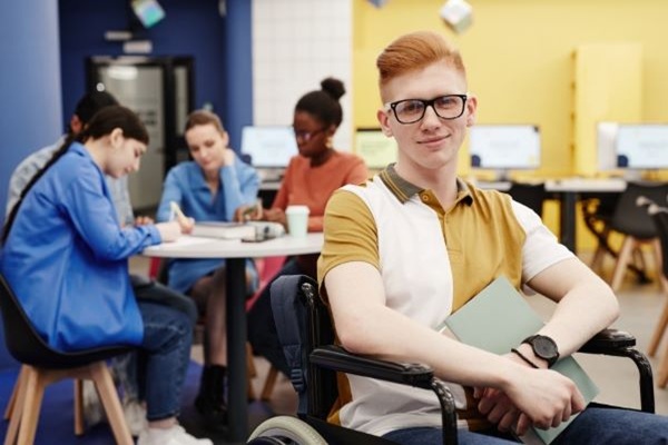 A redheaded light-skinned-young-man using a wheelchair. He is holding a book. There is a group of students studying in the background.
