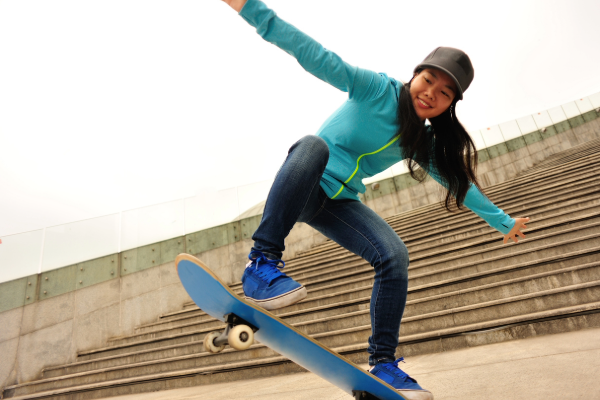 A medium-skinned person wearing a green jacket, blue jeans, and blue skateboarding shoes performing a “kick-flip” on a blue skateboard in front of concrete stairs.