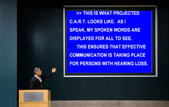 Un hombre trajeado está en un podio y gesticula en dirección hacia una pantalla azul grande detrás de él. En la misma, se muestra CART para una persona con pérdida auditiva. Las palabras en la pantalla son "Así es como CART se ve cuando se proyecta. Mientras hablo, mis palabras se muestran para que todos las puedan ver. Esto garantiza que se está realizando una comunicación efectiva para las personas con pérdida auditiva". Imagen cortesía de Caption Crew.