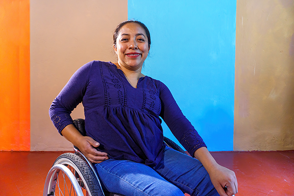 Una mujer de piel morena en silla de ruedas frente a un fondo marrón, naranja y azul. Tiene cabello castaño y lleva una camisa morada con jeans azules.