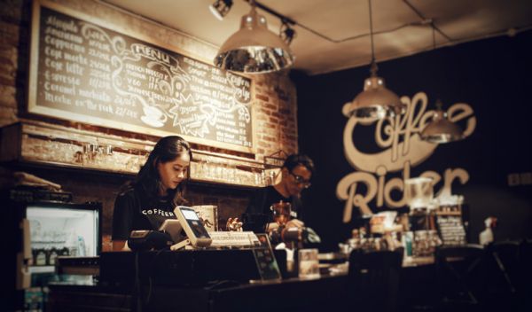 In a dark, cozy coffee shop with items on a chalk board, a female barista is behind the register while a male barista is preparing a drink. This image is for illustrative purposes only and does not depict individuals in the story.