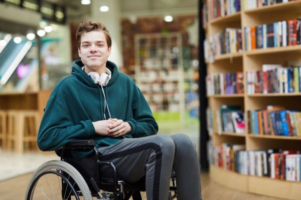 A smiling light-skinned male wheelchair user in a school library.