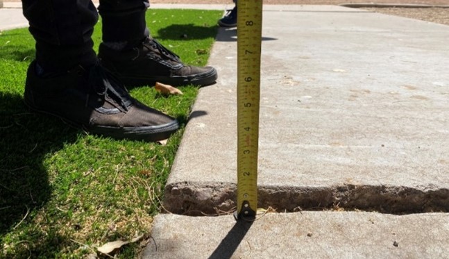 Photograph of someone measuring a cracked sidewalk using a tape measure. The sidewalk is cracked and a portion has risen to form a potential obstacle.