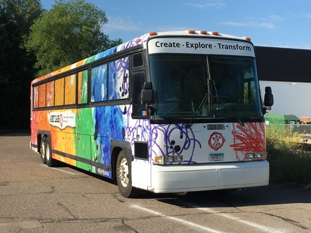 A parked colorful bus with red, orange, green, yellow, and blue splashes of colors. On the top of the windshield it reads, Create - Explore - Transform.