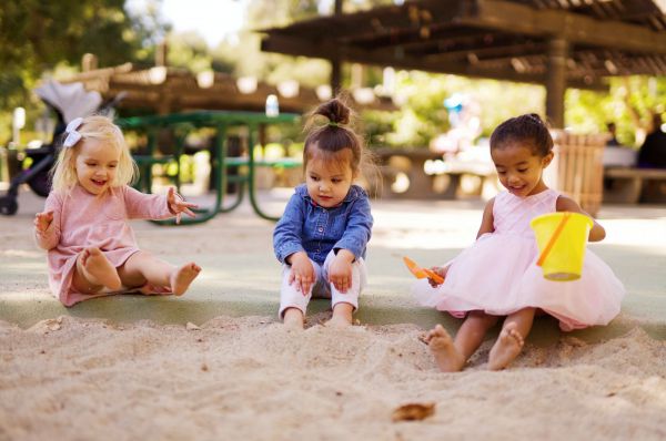 Three young girls playing in a sandbox, one with light skin tone, one with medium skin tone, and one with dark skin tone.