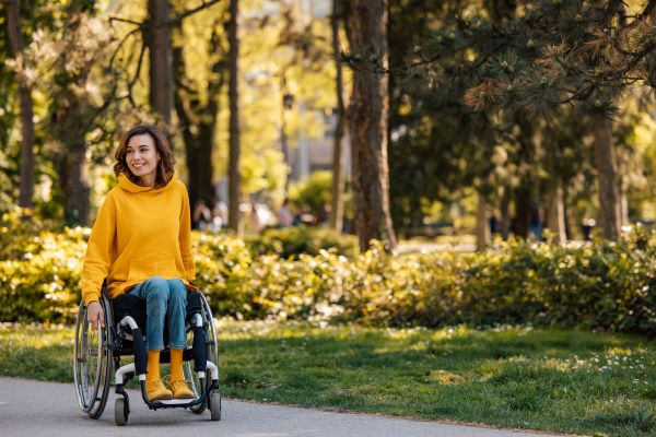 A light-skinned-woman in a bright marigold hoodie and matching shoes uses a wheelchair on an accessible trail in a park. 