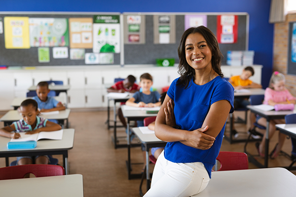 A medium-dark-skinned female teacher smiling with her arms crossed as she leans against a desk at the front of a classroom. The students behind her are working diligently from their workbooks.
