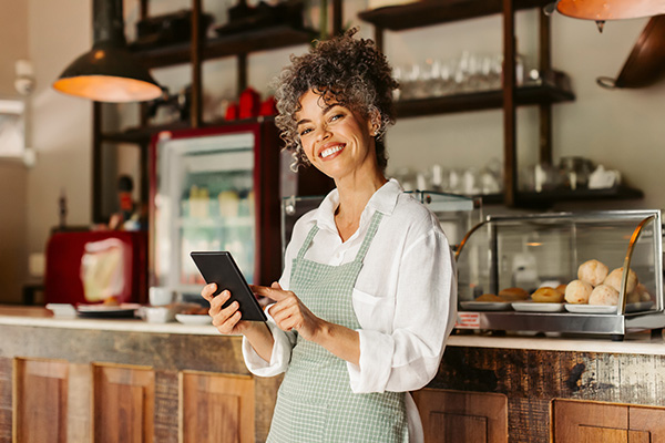 A medium dark-skinned woman with brown curly hair smiling in her coffee shop holding a digital tablet. She is wearing a long sleeve white shirt with a green and white striped apron.