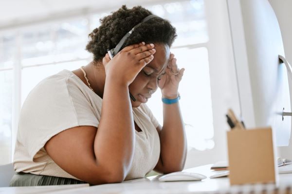 A medium-dark-skinned-woman working in an office. She is wearing a headset and holding her head in her hands in a way that indicates she has a headache.