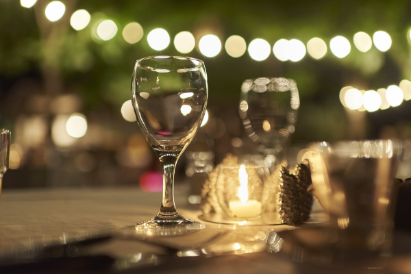 A nighttime outdoor celebration at a restaurant. There are string lights in the background, a table with glasses and a candle. 