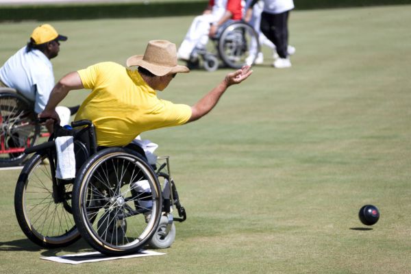 A man wearing a yellow shirt and a tan cowboy hat leans forward in a wheelchair, throwing a large, black ball across a lawn bowling playing field.