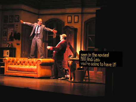 On the left side of a stage, a man stands on the back of a couch and gestures wildly to another man looking up at him.  On the right side of the stage, displayed on a short black screen, closed captioning is provided for an audience at a live theater show.