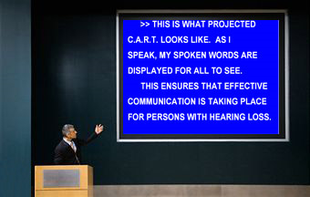 A man in a suit stands at a podium and gestures to a large blue screen behind him.  On it, CART is provided for an individual with hearing loss.  The words on the screen are "This is what projected CART looks like. As I speak, my spoken words are displayed for all to see. This ensures that effective communication is taking place for persons with hearing loss."Image courtesy of Caption Crew.