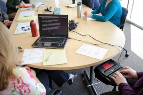 Several people are sitting around a table.  At one end of the table, CART is being provided for an individual with hearing loss on the individual's personal laptop at a work meeting.