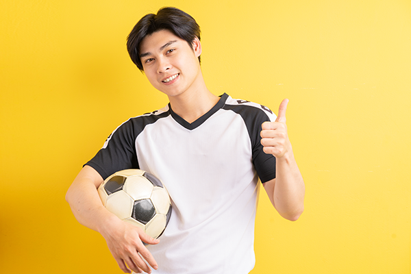 A medium-skinned boy with dark-colored hair is holding a soccer ball in front of a yellow wall. He is wearing a black and white jersey and smiling with a thumbs-up.