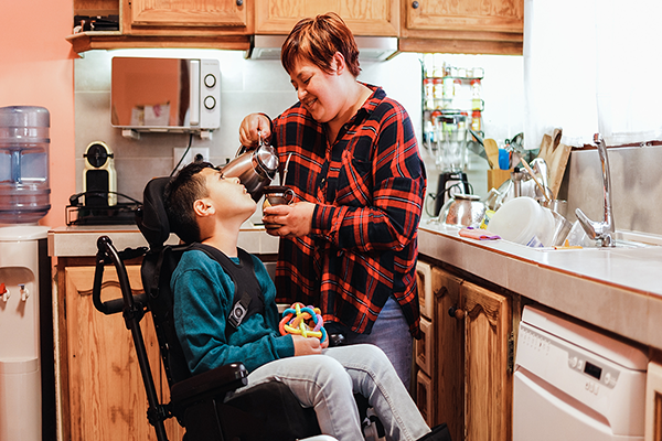 A medium-skinned mother pouring a drink for her son in a wheelchair. She is standing in her kitchen while smiling, wearing a plaid shirt.