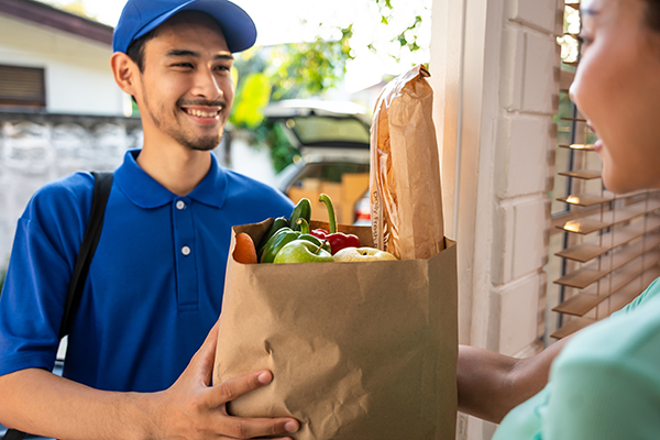 A light-medium-skinned man with dark brown hair hands a medium-skinned woman a bag of groceries.
