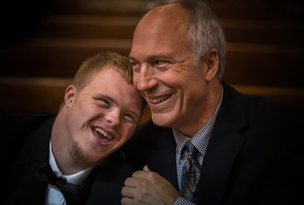 A young man with down syndrome hugs and laughs with an older gentleman: his father. To protect the privacy of individuals involved, people represented on this photograph are not the ones depicted in the story